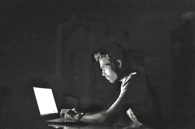 A person sitting in a dark room has their face illuminated by a laptop screen. They are typing intently.