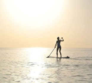 A photo of a person paddleboarding far out into the water, the sun nearing the horizon and no land in sight.