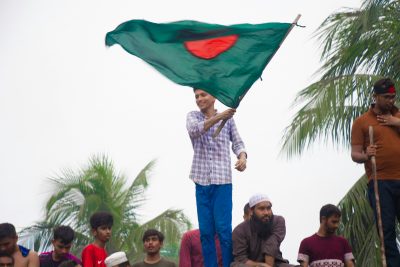 A person in blue pants holds Bangladesh’s flag. People can be seen in the background behind the person waving the country’s green and red flag