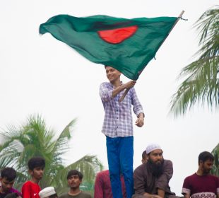 A person in blue pants holds Bangladesh’s flag. People can be seen in the background behind the person waving the country’s green and red flag
