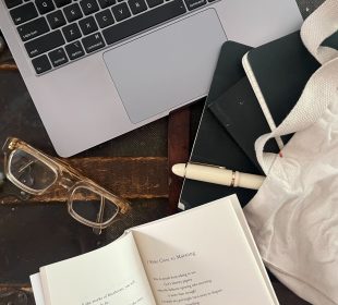 A photo of a laptop, a pair of glasses, an open poem book, and a white tote bag with three black notebooks and a white pen partially inside it, all sitting on what seems to be a vintage chest.
