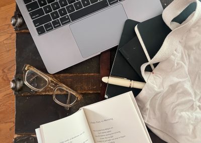 A photo of a laptop, a pair of glasses, an open poem book, and a white tote bag with three black notebooks and a white pen partially inside it, all sitting on what seems to be a vintage chest.