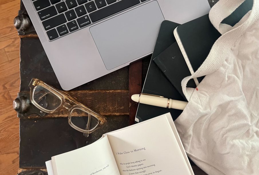 A photo of a laptop, a pair of glasses, an open poem book, and a white tote bag with three black notebooks and a white pen partially inside it, all sitting on what seems to be a vintage chest.