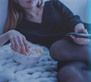 A person with long hair is photographed laying on their side, eating popcorn out of a bowl with one hand and a TV remote in the other.