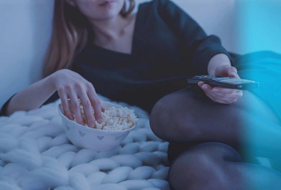 A person with long hair is photographed laying on their side, eating popcorn out of a bowl with one hand and a TV remote in the other.