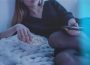 A person with long hair is photographed laying on their side, eating popcorn out of a bowl with one hand and a TV remote in the other.