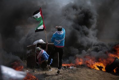 Three people are photographed in front of flames. Two hide in front of a box-like structure, and one is waving a tattered Palestinian flag.