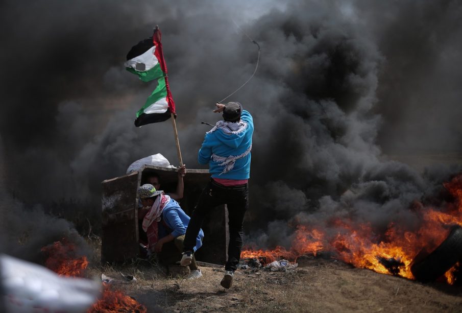 Three people are photographed in front of flames. Two hide in front of a box-like structure, and one is waving a tattered Palestinian flag.
