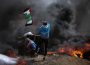 Three people are photographed in front of flames. Two hide in front of a box-like structure, and one is waving a tattered Palestinian flag.