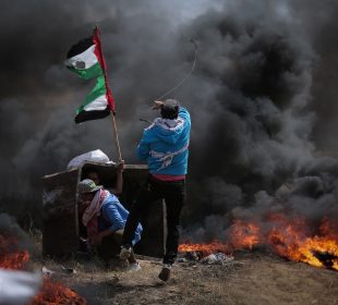 Three people are photographed in front of flames. Two hide in front of a box-like structure, and one is waving a tattered Palestinian flag.