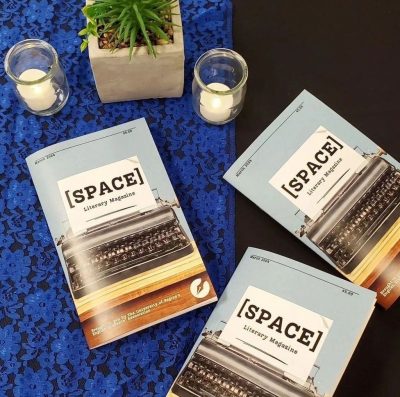 Three [SPACE] magazines laid on a table. A blue lace tablecloth, decorative plant, and two candles are also on the table.