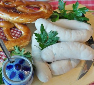 White sausages and golden pretzels displayed on a wooden board on a red and white checkered tablecloth. Also included is a blue lidded pot, a serrated knife, and a threek.