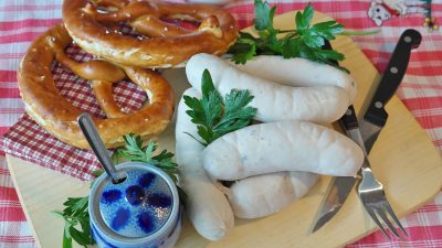 White sausages and golden pretzels displayed on a wooden board on a red and white checkered tablecloth. Also included is a blue lidded pot, a serrated knife, and a threek.