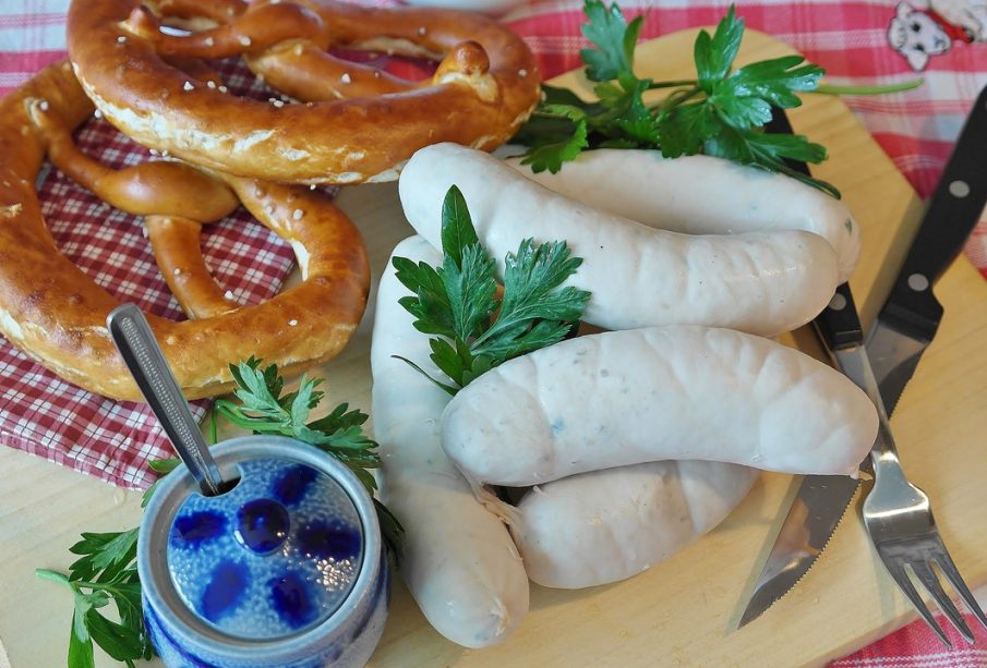 White sausages and golden pretzels displayed on a wooden board on a red and white checkered tablecloth. Also included is a blue lidded pot, a serrated knife, and a threek.