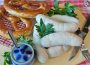 White sausages and golden pretzels displayed on a wooden board on a red and white checkered tablecloth. Also included is a blue lidded pot, a serrated knife, and a threek.