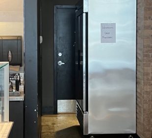 A photo showing a fridge, half-blocking the entrance to the public washroom.