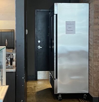 A photo showing a fridge, half-blocking the entrance to the public washroom.