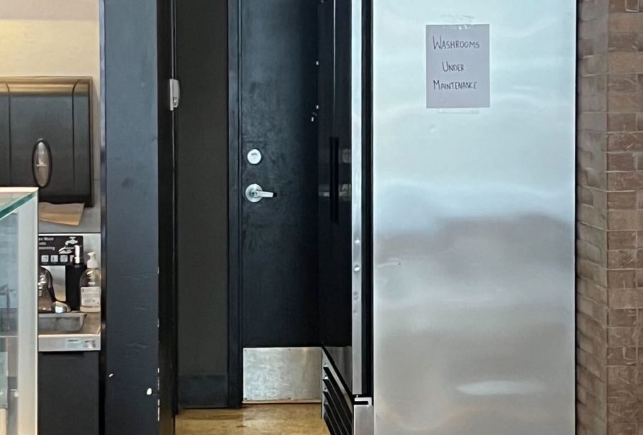 A photo showing a fridge, half-blocking the entrance to the public washroom.