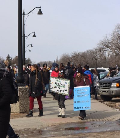 A crowd of teachers picket at the Saskatchewan legislature earlier this year. Two signs are visible, one reads, “SASK STUDENTS AND TEACHERS DESERVE BETTER!”