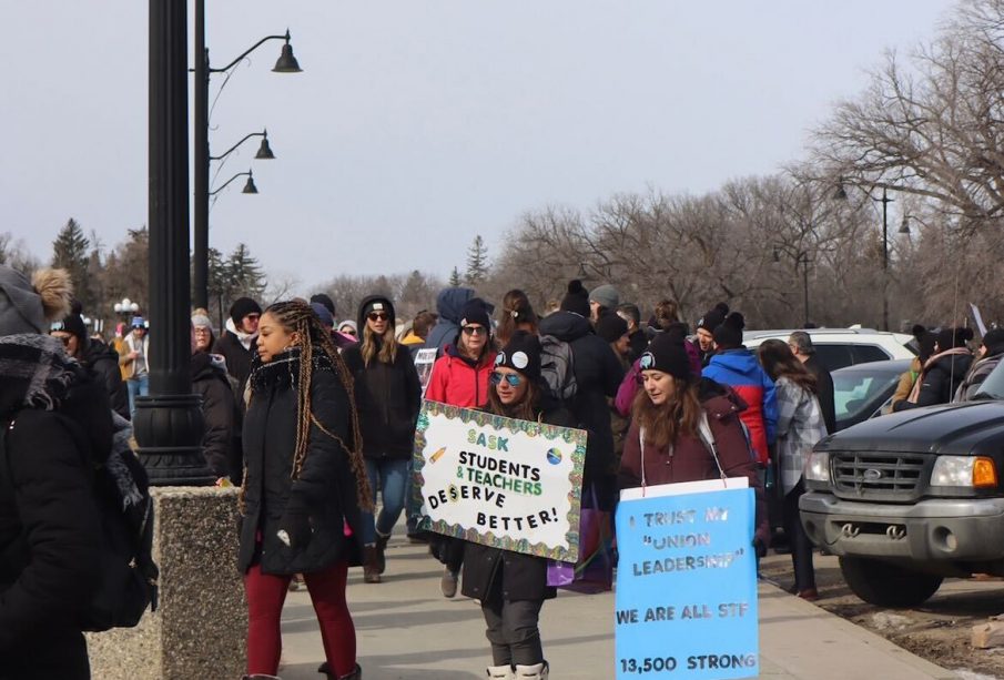 A crowd of teachers picket at the Saskatchewan legislature earlier this year. Two signs are visible, one reads, “SASK STUDENTS AND TEACHERS DESERVE BETTER!”