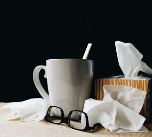 A mug alongside many crumpled tissues and a tissue box. A black pair of glasses rest beside them.