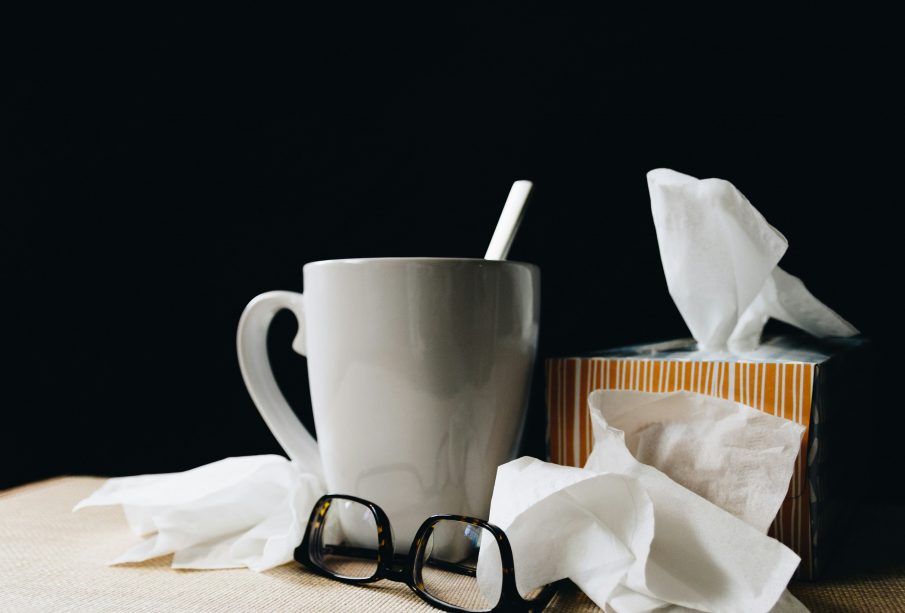 A mug alongside many crumpled tissues and a tissue box. A black pair of glasses rest beside them.