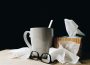 A mug alongside many crumpled tissues and a tissue box. A black pair of glasses rest beside them.