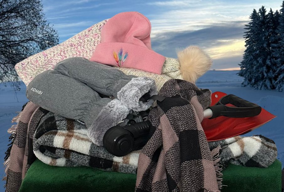 A pile of winter scarves, hats, gloves, and a snow-shovel sit in front of a snow-covered prairie landscape.