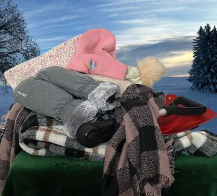A pile of winter scarves, hats, gloves, and a snow-shovel sit in front of a snow-covered prairie landscape.