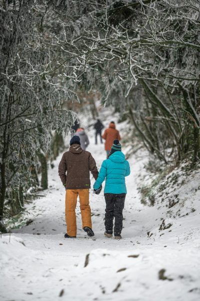 A couple in winter gear walking down in a path in snowy woods, more people ahead.
