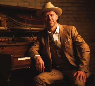 A man in a beige suit and hat sits with his right elbow on a piano, half-smiling.