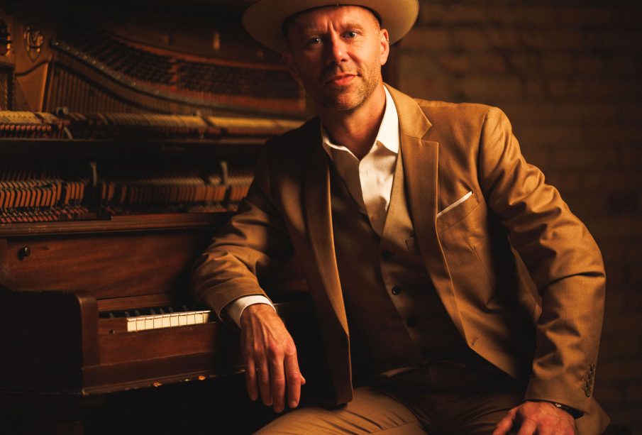 A man in a beige suit and hat sits with his right elbow on a piano, half-smiling.