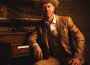 A man in a beige suit and hat sits with his right elbow on a piano, half-smiling.