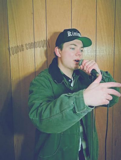 Brett Rybinski in front of a wood-paneled wall with the words “Young Corduroy” across it.