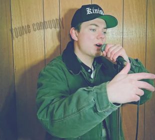 Brett Rybinski in front of a wood-paneled wall with the words “Young Corduroy” across it.