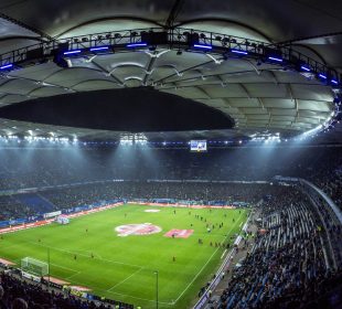 Full shot of a soccer field and surrounding stadium seats full of fans, with players scattered on the field