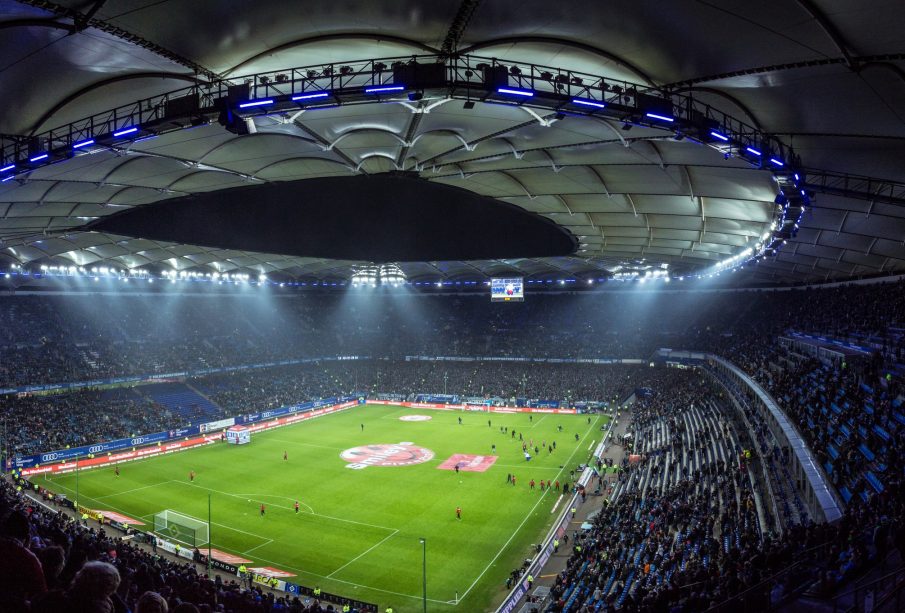Full shot of a soccer field and surrounding stadium seats full of fans, with players scattered on the field