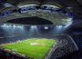 Full shot of a soccer field and surrounding stadium seats full of fans, with players scattered on the field