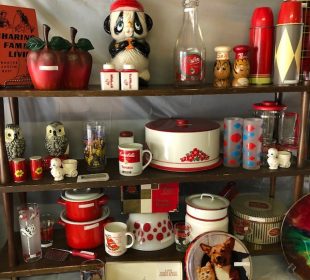 Three shelves with strategically displayed items one could find at the vintage market all following a red and white colour scheme, items such as: metal apples, a panda bear, a bottle, thermoses, owls, salt and pepper shakers, a Campbell’s soup mug, some pots and bowls, and a plate with a corgi and a kitten.