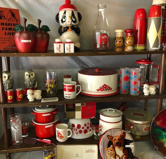 Three shelves with strategically displayed items one could find at the vintage market all following a red and white colour scheme, items such as: metal apples, a panda bear, a bottle, thermoses, owls, salt and pepper shakers, a Campbell’s soup mug, some pots and bowls, and a plate with a corgi and a kitten.