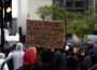 Black person in red hood holds sign that reads “It’s a privilege to learn about racism instead of experiencing it!!!”