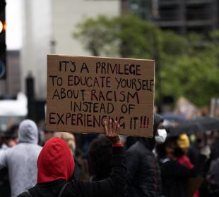 Black person in red hood holds sign that reads “It’s a privilege to learn about racism instead of experiencing it!!!”