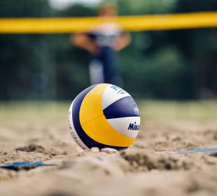 Close up of a blue, yellow, and white volleyball placed in the sand with blurry players and net in the background