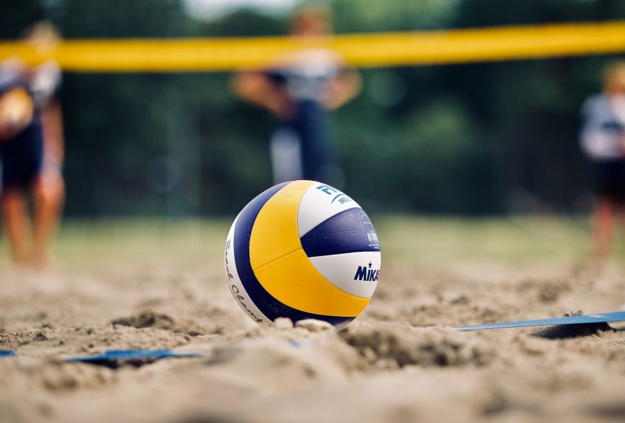 Close up of a blue, yellow, and white volleyball placed in the sand with blurry players and net in the background