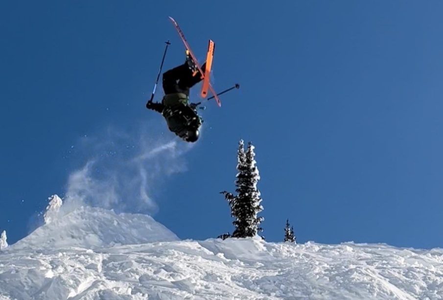 A skier jumps off a ramp, doing a flip mid-air with the orange bottoms of their skis in full view.