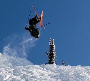 A skier jumps off a ramp, doing a flip mid-air with the orange bottoms of their skis in full view.