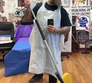 This is a selfie of the owner in his shop. He is wearing a white apron, his arms are covered in tattoos, he has a medium beard and glasses. He also is holding a mop in the picture, and the walls in the background are covered in collected art pieces.