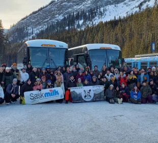 A group photo of students in Banff on the ski and board trip, posing in front of their charter buses and a tree-covered mountain. 