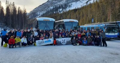 A group photo of students in Banff on the ski and board trip, posing in front of their charter buses and a tree-covered mountain.
