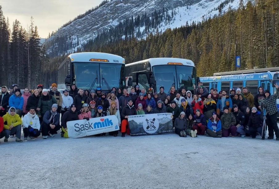 A group photo of students in Banff on the ski and board trip, posing in front of their charter buses and a tree-covered mountain. 